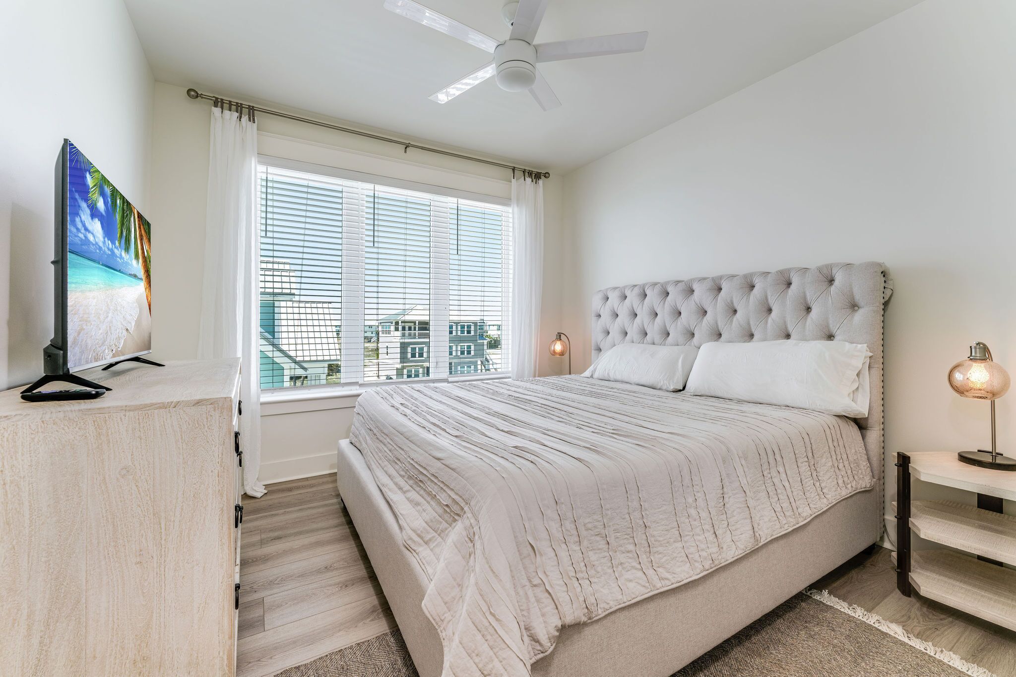 A bedroom with a large bed, a ceiling fan, a TV on a wooden cabinet, and a window with blinds.