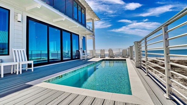 A modern outdoor swimming pool with a wooden deck, large glass windows, and a view of the ocean.