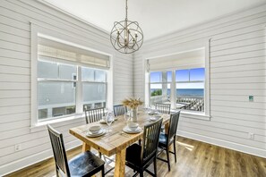 A dining room with a wooden table set for six, large windows with blinds, and a view of the ocean.