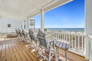 A wooden deck with Adirondack chairs and a table, overlooking a beach with a clear view of the ocean.
