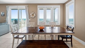 Un comedor con mesa de madera, sillas blancas y un tazón. Hay ventanas con persianas, vista al océano y una pared de color claro.
