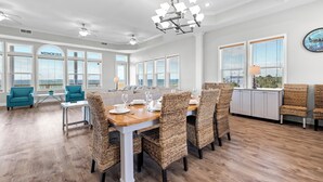A spacious dining area with a wooden table, wicker chairs, and a view of the ocean through large windows. - Memory Maker: Beachfront, Fire Pit, Sleeps 18! (St. Joe Beach)