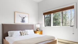 A bedroom with a wooden bed, white bedding, a framed artwork of a bull, a bedside table with a lamp, and a window with a view of trees. - Outpost: Balsam Lodge B7 (Wilson)