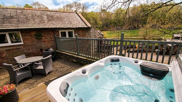 Un bain à remous décoré avec un motif d’arbre, une terrasse en bois, des meubles en osier et un bâtiment en pierre entouré d’une clôture.