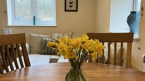 Un coin repas avec une table en bois, des chaises, un vase rempli de fleurs jaunes, un canapé avec des coussins à rayures et une fenêtre donnant sur des arbres.