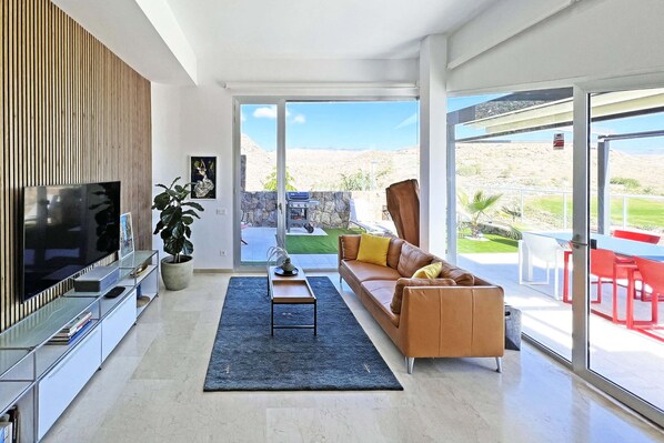 A modern living room with a flat-screen TV, a sofa, a coffee table, and a large window offering a view of a landscaped outdoor area.