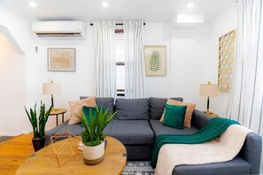 Uma sala de estar moderna com um sofá cinza, almofadas verdes e bege, uma mesa de centro de madeira e duas mesas laterais com abajures.