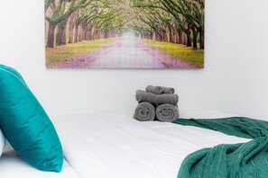 Uma cama com um quadro de uma estrada bordeada por árvores, uma pilha de toalhas enroladas e um travesseiro azul-turquesa.