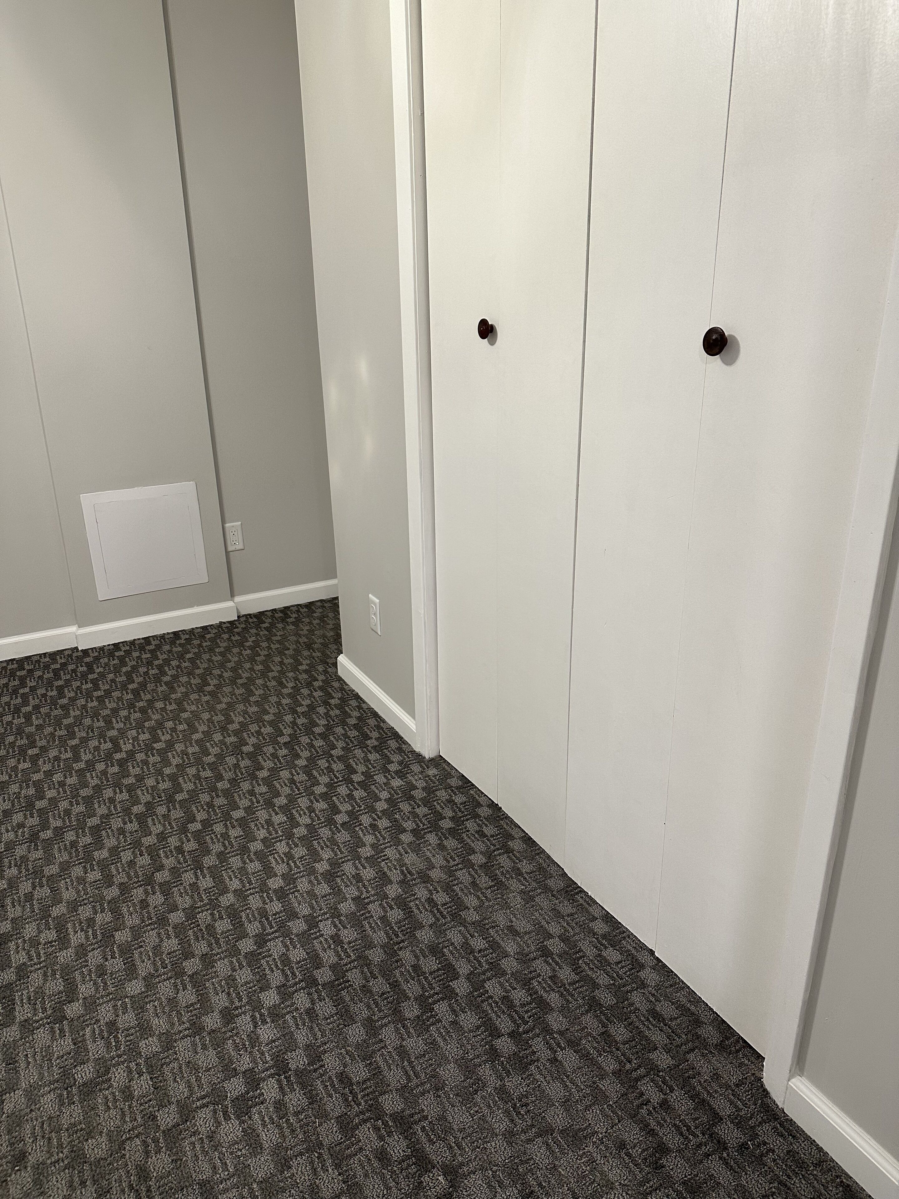 Une pièce avec un tapis à motifs, une porte blanche dotée de deux poignées rondes et une petite grille d’aération sur le mur.