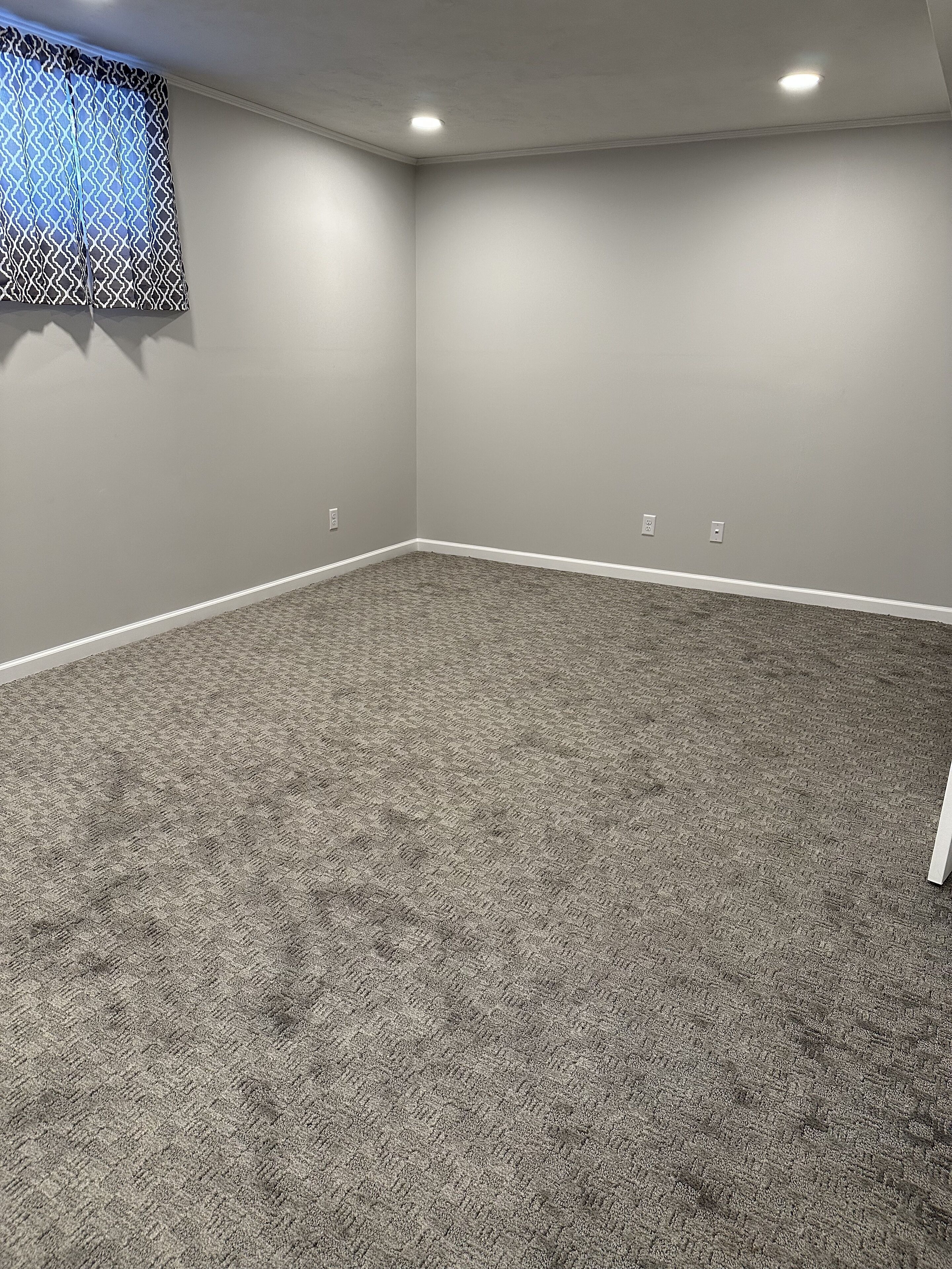 Een kamer met vloerbedekking, witte muren en een raam met een geometrisch patroon.