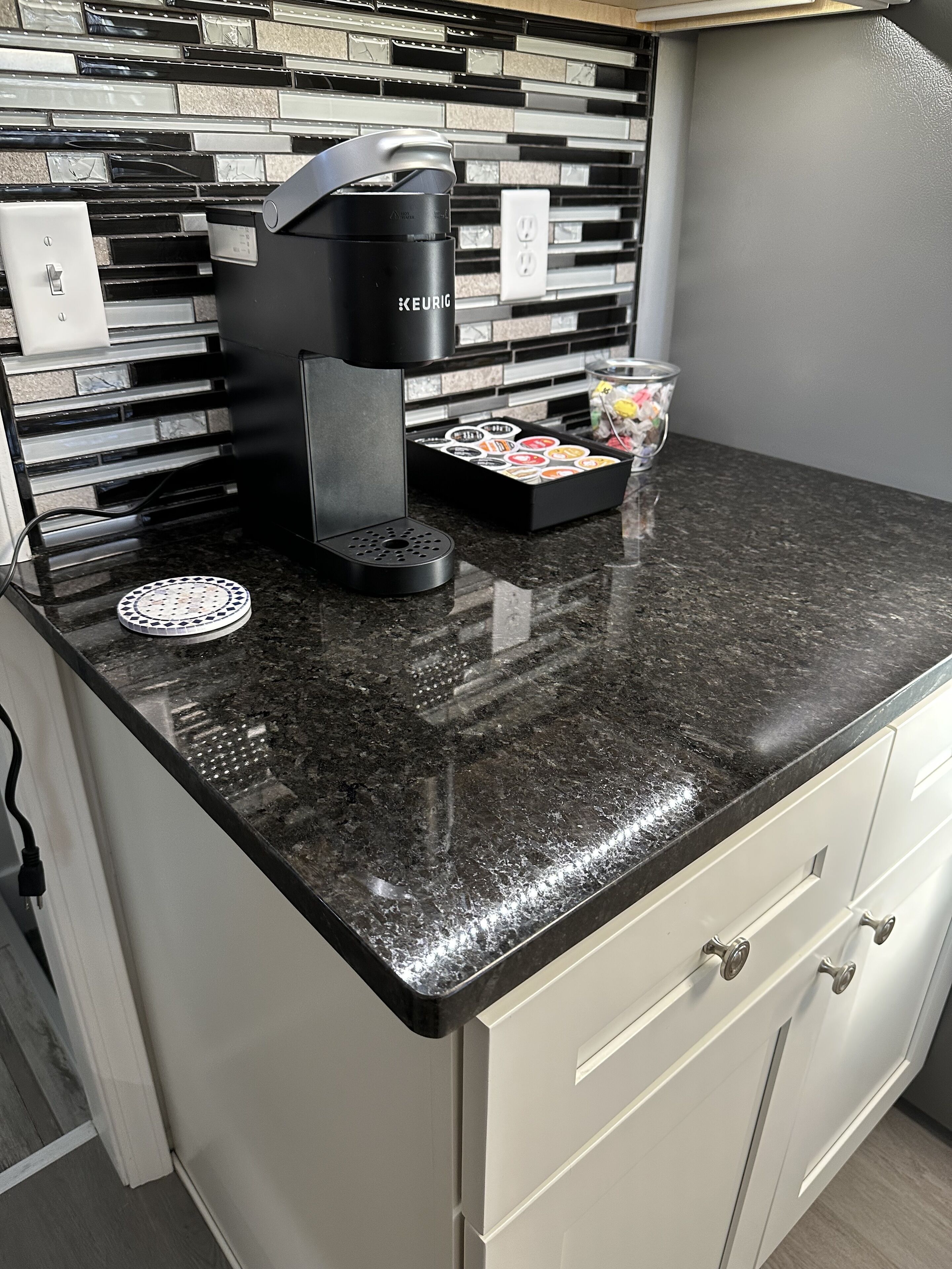 A kitchen with a black countertop, a Keurig coffee maker, a coffee pod holder, and a trash can.