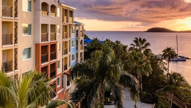 Un hôtel à plusieurs étages doté de balcons donnant sur un plan d’eau et bordé de palmiers.