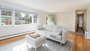 A spacious living room with a white sectional sofa, a matching ottoman, and a large window providing natural light. - Recently Updated, One Level, Attached Garage, Peaceful, Zen-Minimalist Space.  (Avon)
