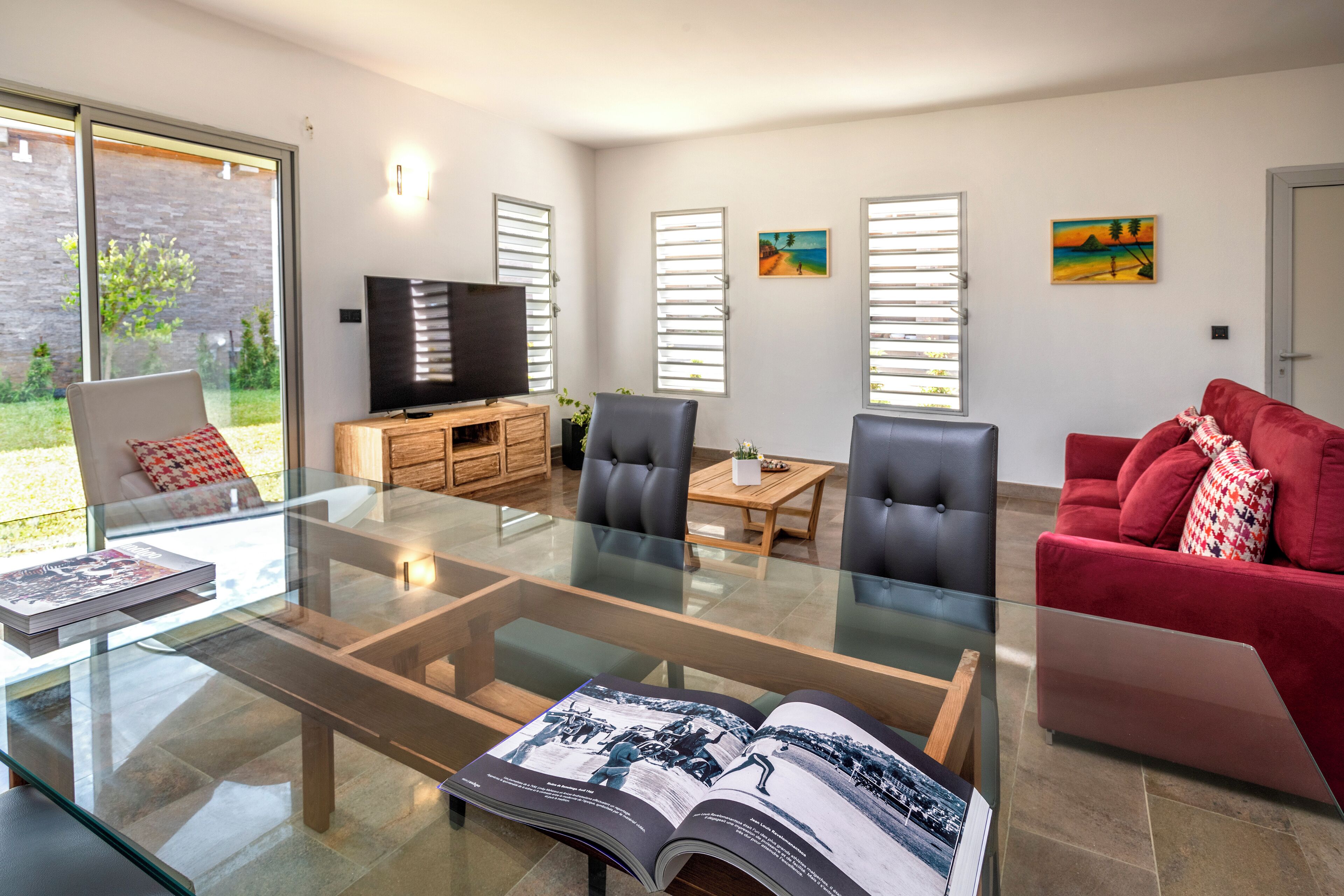 A modern living room with a glass coffee table, leather chairs, a red sofa, and a television on a wooden cabinet.