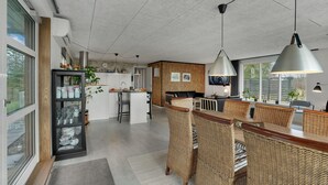 A modern kitchen with a central island, a dining area with wicker chairs, and large windows providing natural light. - 3 bedroom cozy home in Thyholm (Thyholm)