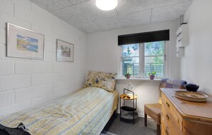 Une petite pièce bien éclairée, avec un lit, une petite table, une commode et une fenêtre ornée de plantes.