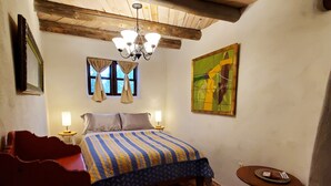 Un dormitorio con una cama, una mesita de noche, una pequeña mesa, una silla, una alfombra, un cuadro y una ventana con cortinas.