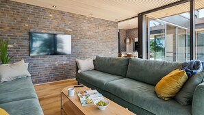 A modern living room with a brick wall, a large flat-screen TV, a green sofa, a wooden coffee table with magazines and a bowl of fruit, and a large window offering a view outside. - Gorgeous home in Ebeltoft with kitchen (Ebeltoft)