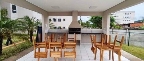 Un patio al aire libre con suelo de baldosas, sillas de madera y zona de barbacoa.