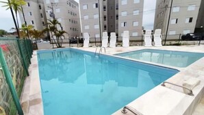 A swimming pool with a tiled edge and a metal handrail, surrounded by lounge chairs and palm trees, with a building in the background.
