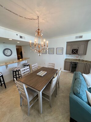 A dining area with a wooden table and chairs, a chandelier, and a kitchen with cabinets and a refrigerator in the background.