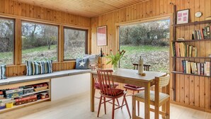 A cozy cabin interior with a wooden interior, a dining table, chairs, a bookshelf, and a window seat overlooking a wooded area. - 2 bedroom amazing home in Jægerspris (Jægerspris)