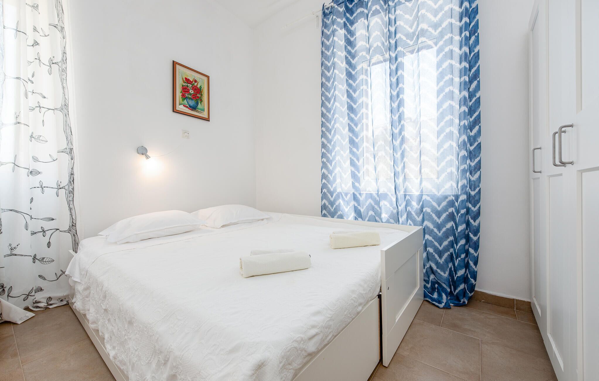 Un dormitorio con cama, cortinas blancas, una lámpara fijada a la pared y un cuadro pequeño enmarcado.