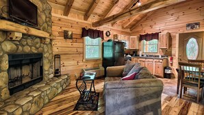 A cozy cabin interior with a stone fireplace, wooden walls, a dining area, and a kitchen.