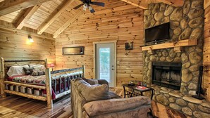 A log cabin interior with a bed, a sofa, a stone fireplace, and a ceiling fan.