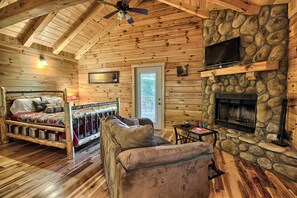 Interior de una cabaña de troncos con una cama, un sofá, una chimenea de piedra y un televisor en la pared.