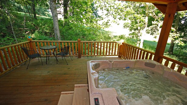 A wooden deck with a hot tub, a table with chairs, and a view of a forested area.