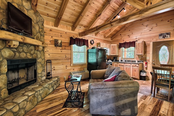 L’intérieur d’un chalet rustique, avec une cheminée en pierre, des murs en bois, un téléviseur, un coin repas et une cuisine.
