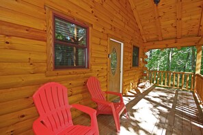 A wooden cabin with two red Adirondack chairs on the porch, a door with a glass panel, and a window with blinds.