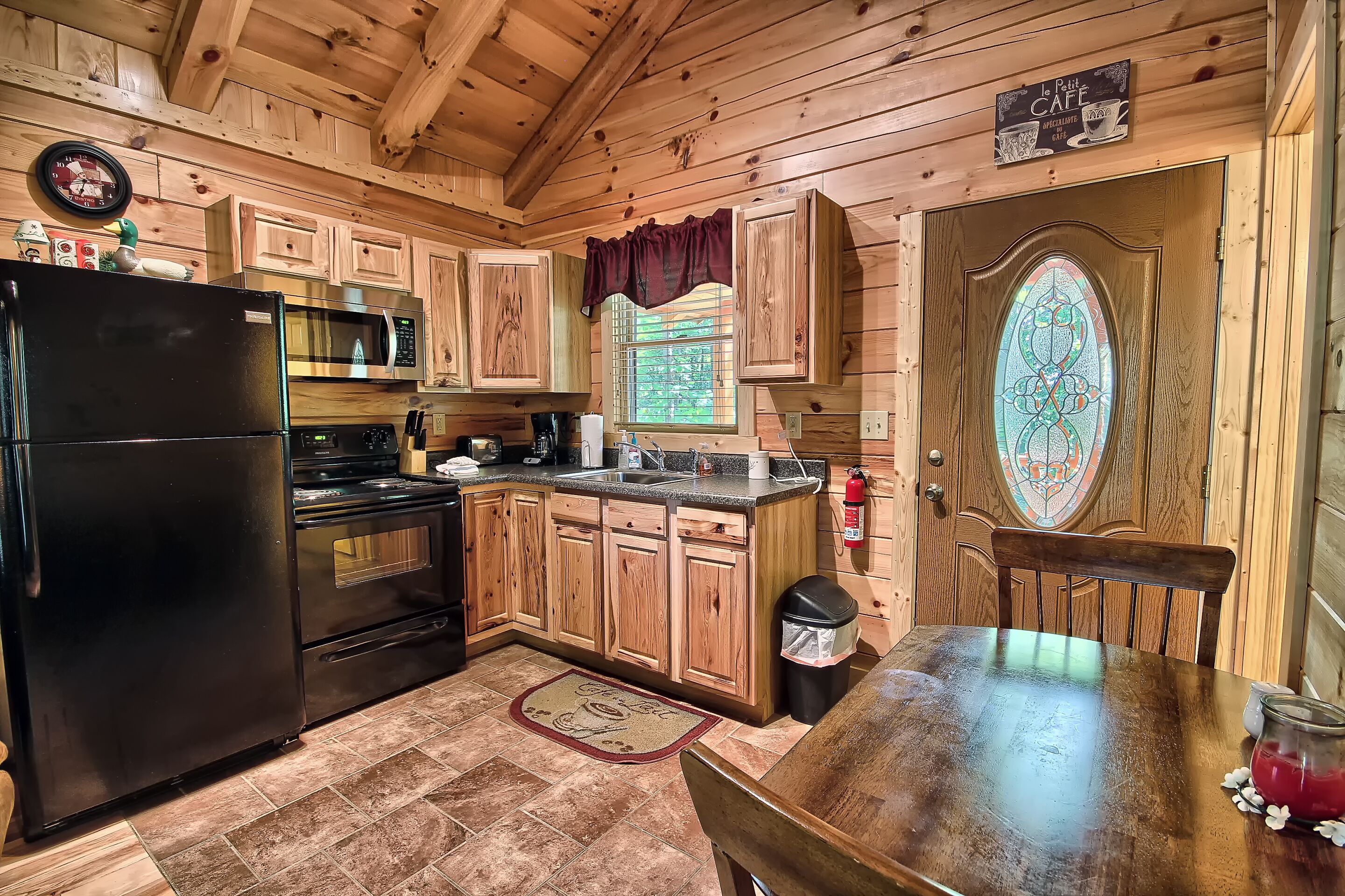 A wooden cabin kitchen with a black refrigerator, stainless steel microwave, and stove, wooden cabinets, a window with a view, and a stained glass door.