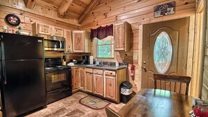 A wooden cabin kitchen with a black refrigerator, stainless steel microwave, and stove, wooden cabinets, a window with a view, and a stained glass door.