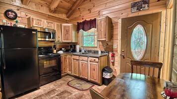 A wooden cabin kitchen with a black refrigerator, stainless steel microwave, and stove, wooden cabinets, a window with a view, and a stained glass door.