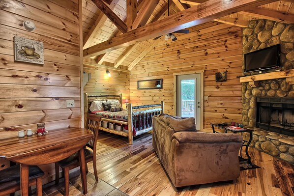 A cozy cabin interior with a wooden floor, a stone fireplace, a television mounted above it, and a bed with a colorful blanket.