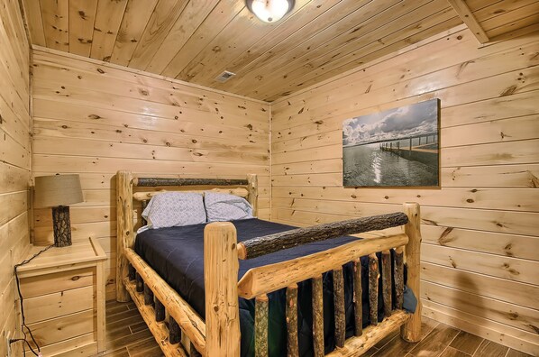 A wooden cabin bedroom with a bed, nightstand, lamp, and a painting of a bridge.