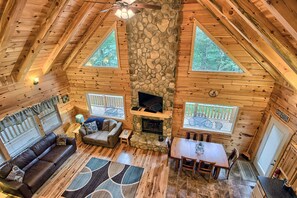 A cozy cabin interior with a stone fireplace, wooden walls, a sofa, a dining table, and two windows with views of trees.