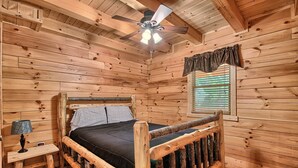 Een slaapkamer in een houten hutje, met een bed, nachtkastje, lamp, plafondventilator en een raam met jaloezieën.