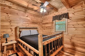 Ein Schlafzimmer in einer Holzhütte mit einem Bett, einem Nachttisch, einer Lampe, einem Deckenventilator und einem Fenster mit Jalousien.