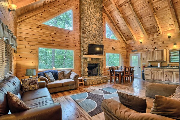 A log cabin interior with a stone fireplace, wooden furniture, and large windows.