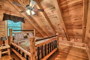 Ein Schlafzimmer einer Holzhütte mit schrägem Dach, einem Bett mit farbenfroher Steppdecke, einem Nachttisch mit Lampe und einem Fenster mit Jalousien.