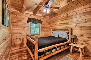 Une chambre à coucher située dans une cabane en bois, comprenant un lit, une table de chevet, un ventilateur de plafond et une fenêtre avec des stores.
