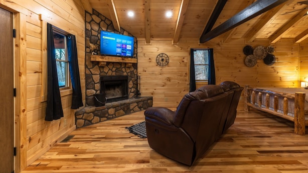 L’intérieur chaleureux d’un chalet avec une cheminée en pierre, des murs en bois, un fauteuil inclinable en cuir et un téléviseur posé sur une étagère en pierre.