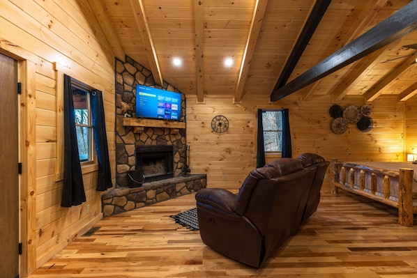 A cozy cabin interior with a stone fireplace, wooden walls, a leather recliner, and a television on a stone shelf.