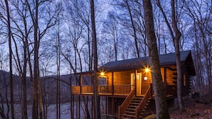 Une cabane dotée d’une terrasse en bois et d’un escalier menant à un deuxième étage, entourée d’arbres et bordée par un plan d’eau.