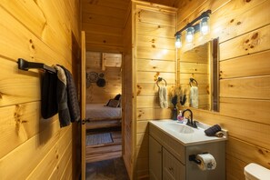 Interior de una cabaña de madera con una puerta de madera que da a un dormitorio, un baño con lavabo y espejo, y un rollo de papel higiénico a la vista.