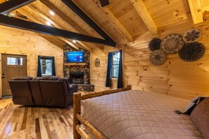Un acogedor interior de cabaña de troncos con una cama, un sofá, una chimenea y un televisor montado en la pared.
