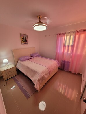 A bedroom with a bed, a nightstand, a lamp, a ceiling fan, and a window with curtains.